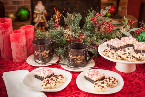 A festive table setting with holiday treats, including cookies and brownies, surrounded by greenery and candles.