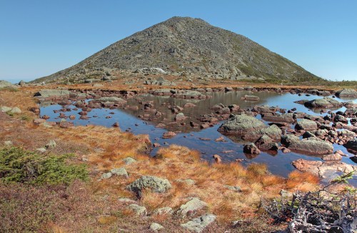 A rocky mountain rises above a serene pond, surrounded by grassy terrain and scattered boulders under a clear blue sky.