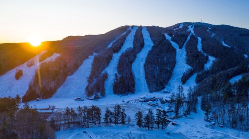 A snowy mountain landscape at sunset, featuring ski slopes and a cluster of buildings at the base.
