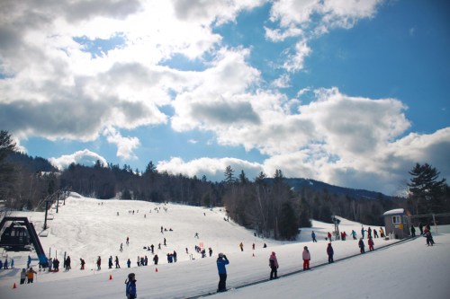 A snowy ski slope with people skiing and snowboarding under a partly cloudy sky. Trees line the background.