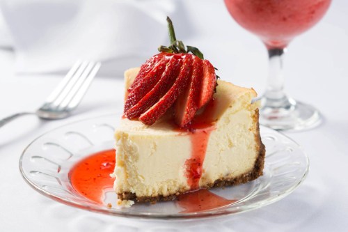 A slice of cheesecake topped with fresh strawberries and strawberry sauce, served on a glass plate with a drink in the background.