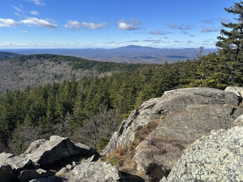 A panoramic view from a rocky outcrop, showcasing lush green forests and distant mountains under a clear blue sky.