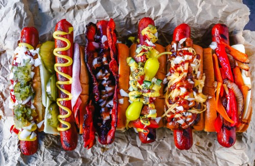A row of colorful hot dogs with various toppings, including mustard, onions, and peppers, on crumpled brown paper.