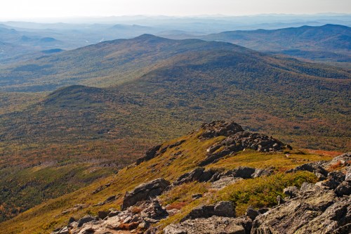 A panoramic view of rolling mountains covered in green and orange foliage under a clear sky. Rocky foreground.