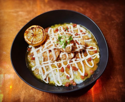 A bowl of creamy risotto topped with shrimp, crispy bacon, drizzled with sauce, and garnished with a grilled lemon.