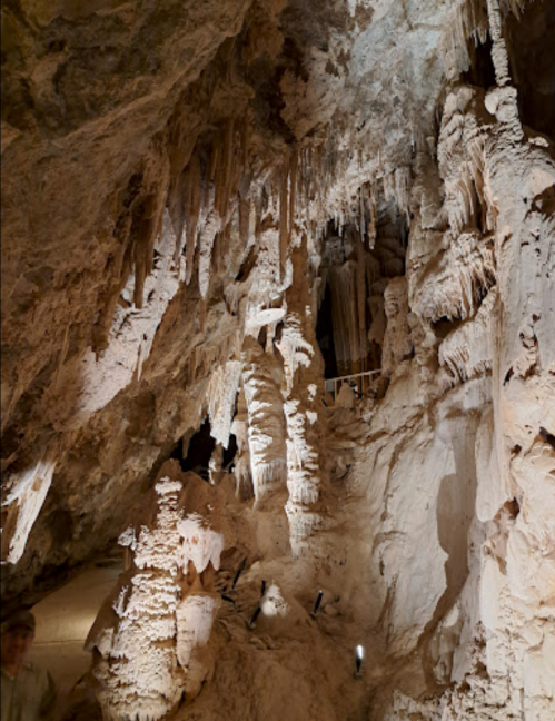 A dimly lit cave interior featuring intricate stalactites and stalagmites, with a person standing in the foreground.