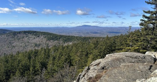A panoramic view of rolling hills and mountains under a blue sky, with a rocky foreground and dense green forest.