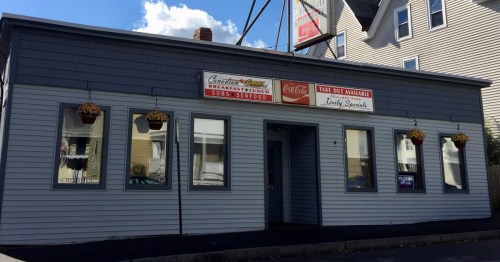 A gray building with a sign for a restaurant offering breakfast, subs, and takeout, featuring hanging flower pots.