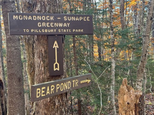 Wooden trail signs in a forest, directing to Monadnock-Sunapee Greenway and Bear Pond Trail. Autumn foliage in background.