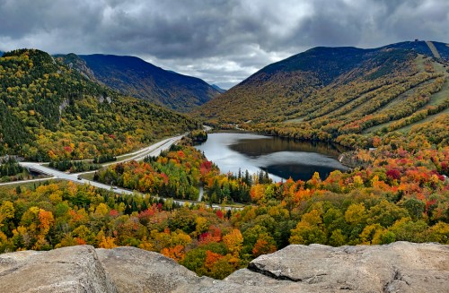 A scenic view of a lake surrounded by colorful autumn foliage and mountains under a cloudy sky.