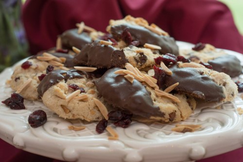 A plate of cookies topped with chocolate, almonds, and dried cranberries, set on a decorative white platter.