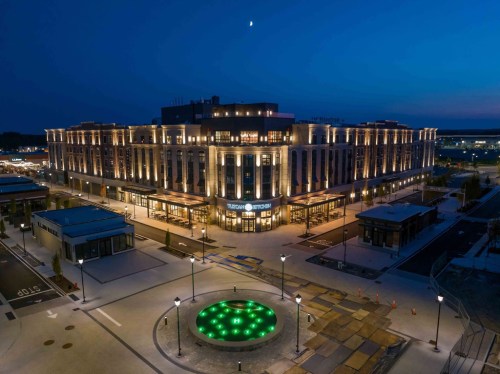 A modern building illuminated at night, featuring a circular green fountain in the foreground and a clear sky above.