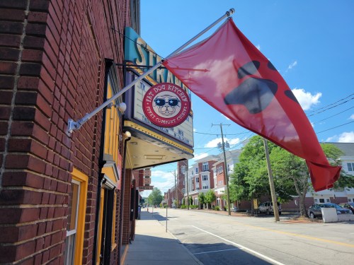 A red flag flutters outside a restaurant with a sign reading "Sita's Dog Kitchen" on a sunny street.