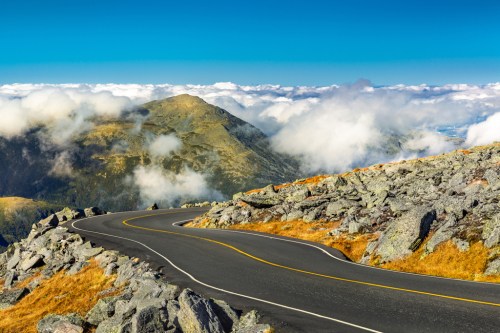 A winding road curves through rocky terrain with mountains and clouds in the background under a clear blue sky.