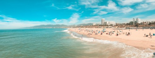 A sunny beach scene with people enjoying the sand and water, framed by mountains and buildings in the background.