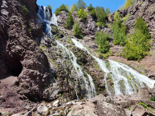 A cascading waterfall flows down rocky cliffs, surrounded by lush greenery and a clear blue sky.