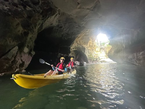 Two people in a yellow kayak explore a cave, with light streaming in from a large opening ahead.