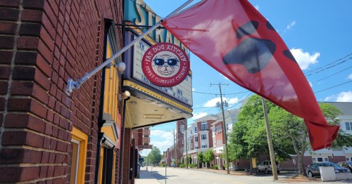 A red flag and sign for "Fat Dog Kitchen" on a brick building, with a clear blue sky and street view in the background.