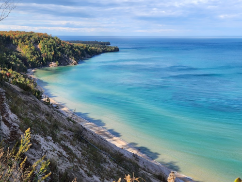 A serene coastal view with turquoise waters, sandy beach, and lush green cliffs under a partly cloudy sky.