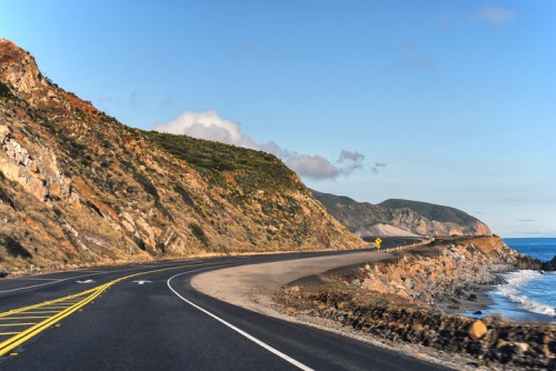 A winding coastal road curves along rocky cliffs with the ocean visible on the right under a clear blue sky.