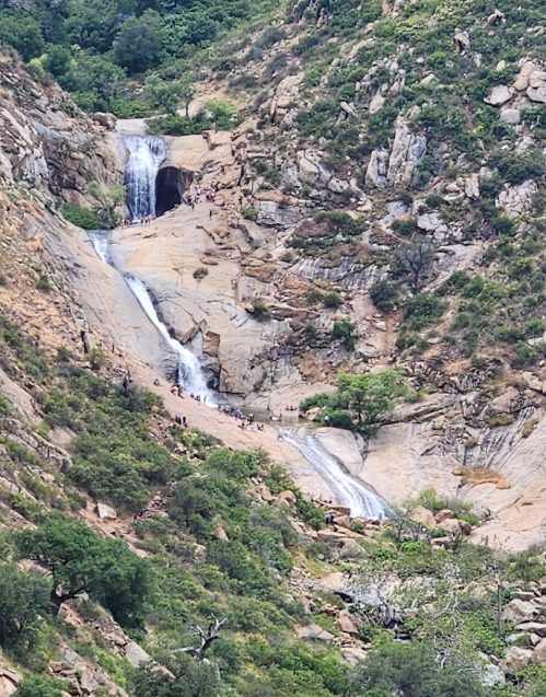 A scenic view of a waterfall cascading down rocky terrain, surrounded by lush greenery and small groups of people.