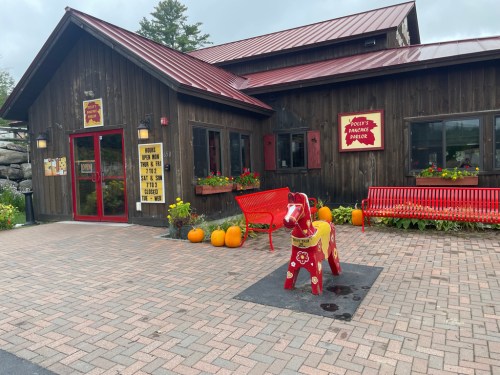 A rustic wooden building with a red roof, featuring a colorful horse statue and pumpkins in front.