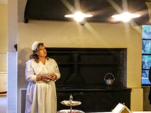 A woman in a vintage dress and apron stands in a kitchen, speaking near a large black stove and a table with treats.