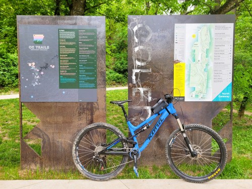 A blue mountain bike parked next to trail maps and information boards in a green park setting.