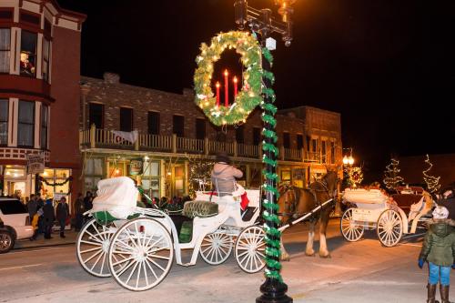 A festive night scene with horse-drawn carriages, holiday decorations, and people enjoying a winter celebration.