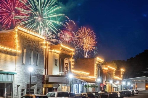 Colorful fireworks illuminate a night sky above a charming, lit-up downtown street with historic buildings.