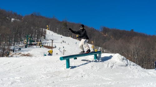 A snowboarder performs a trick on a rail at a snowy ski resort under a clear blue sky.