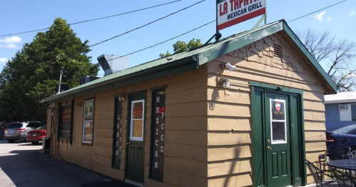 A small, rustic building with a green roof and sign reading "La Tapatia Mexican Grill," surrounded by trees and parked cars.