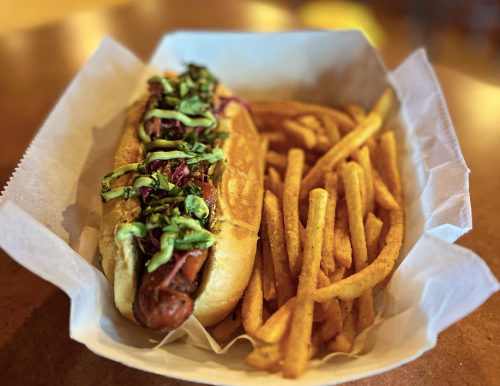 A hot dog topped with slaw in a bun, served with a side of golden, crispy fries in a paper-lined basket.