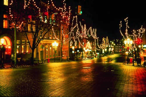 A quiet street at night, lined with buildings adorned with festive lights reflecting on the wet pavement.