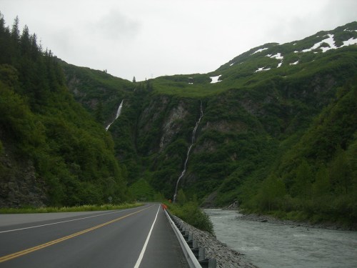 A scenic road alongside a river, with lush green mountains and waterfalls cascading down the cliffs.