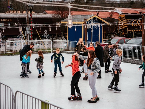 A lively ice skating rink with children and adults skating, surrounded by festive decorations and a train in the background.