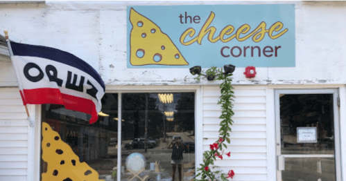 A storefront with a sign reading "The Cheese Corner" and a flag featuring the word "OREZ." Greenery and flowers adorn the entrance.