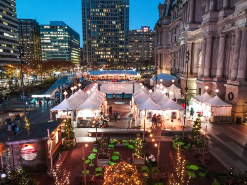 A festive outdoor market with tents, lights, and holiday decorations, set against a city skyline at dusk.