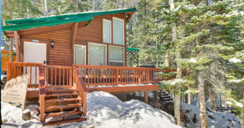 A cozy wooden chalet surrounded by snow-covered trees, featuring a deck and large windows.