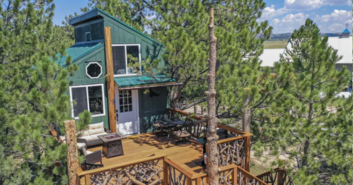 A cozy green cabin nestled among tall pine trees, featuring a wooden deck and outdoor seating.
