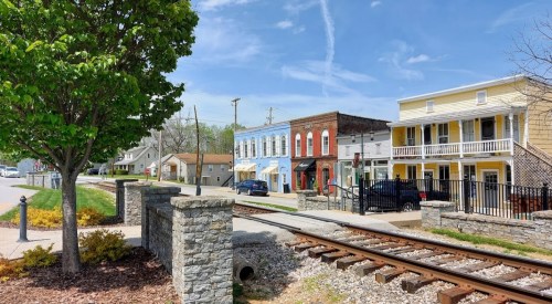 A quaint street scene with colorful buildings, a tree, and railroad tracks under a clear blue sky.