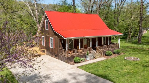 A charming brown house with a red roof, surrounded by greenery and a concrete driveway.