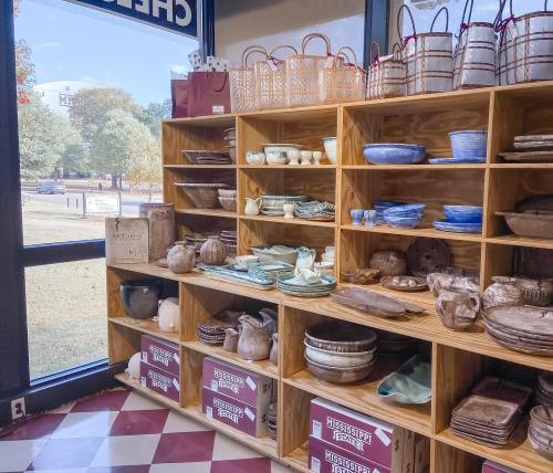 Wooden shelves display various pottery and kitchenware, with a view of trees and a street outside the window.