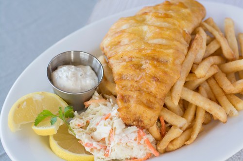 A plate of golden-brown fried fish with fries, coleslaw, lemon wedges, and a small cup of tartar sauce.