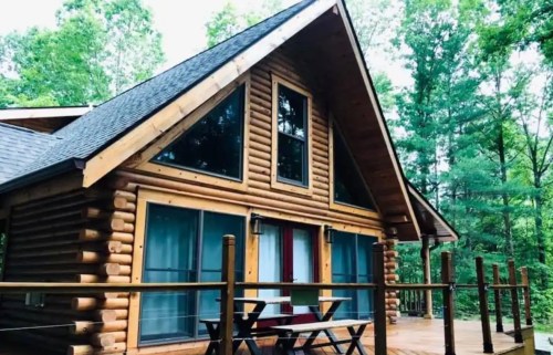 A cozy log cabin with large windows, surrounded by trees, featuring a deck and outdoor seating.