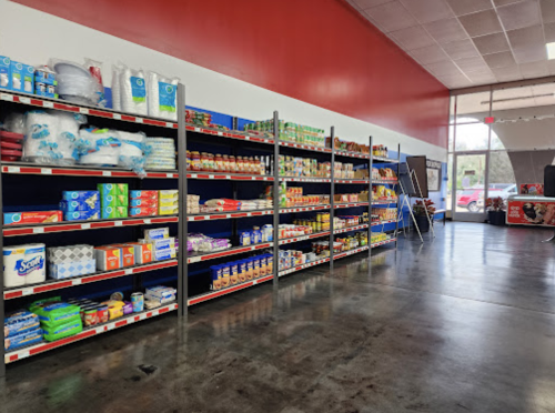 A well-stocked grocery store aisle with shelves of various food products and supplies against a red wall.
