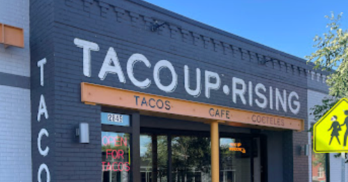 Exterior of Tacoup Rising, a taco cafe with a sign that reads "OPEN FOR TACOS" and a bright blue sky above.