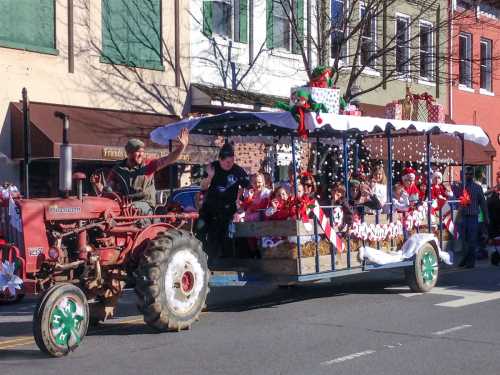 A festive parade float decorated for the holidays, pulled by a tractor, with children and adults waving joyfully.