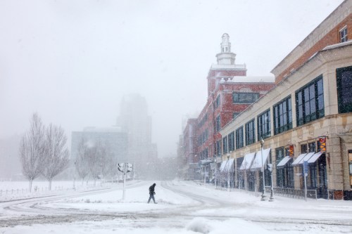 A snowy street scene with a lone pedestrian walking past buildings in a winter storm. Visibility is low due to heavy snowfall.