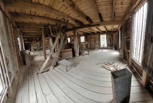 Interior of an old wooden building with exposed beams, wooden floors, and scattered debris. Natural light through windows.